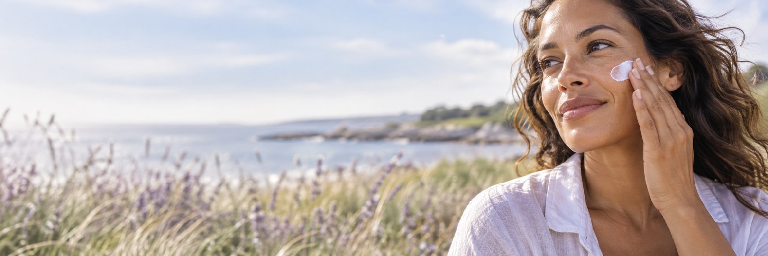 Woman applying sunscreen to her cheek beside a coastal landscape.